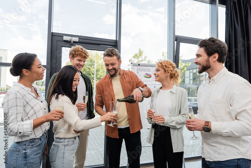 Photography cheerful man pouring champagne near multicultural friends during corporate party