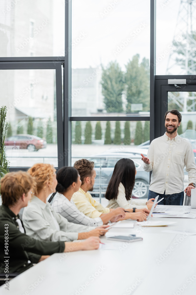 Fototapeta premium positive bearded businessman pointing with hand while talking to multiethnic colleagues
