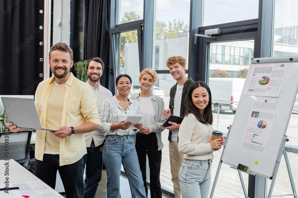 cheerful multiethnic corporate team looking at camera near flip chart with infographics cheerful multiethnic corporate team looking at camera near flip chart with infographics