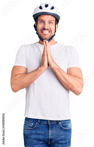 Young handsome man wearing bike helmet praying with hands together asking for forgiveness smiling confident.