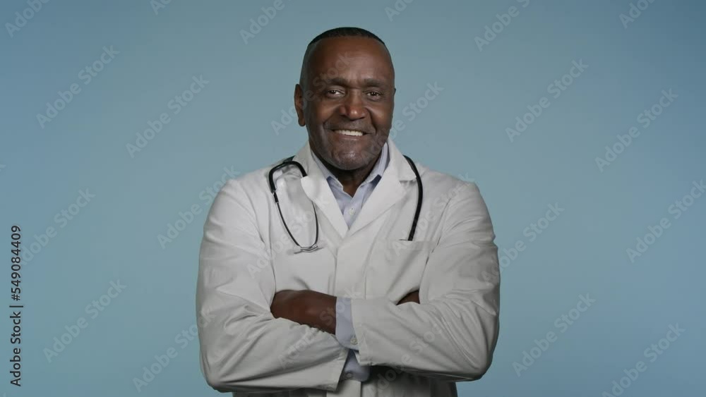 Mature confident happy doctor standing in gray background studio with arms crossed glad smiling african american man professional therapist physician in medical gown posing indoors looking at camera Mature confident happy doctor standing in gray background studio with arms crossed glad smiling african american man professional therapist physician in medical gown posing indoors looking at camera