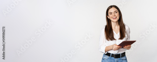 Smiling modern woman standing with digital tablet, laughing and looking happy, working, posing against white studio background photo