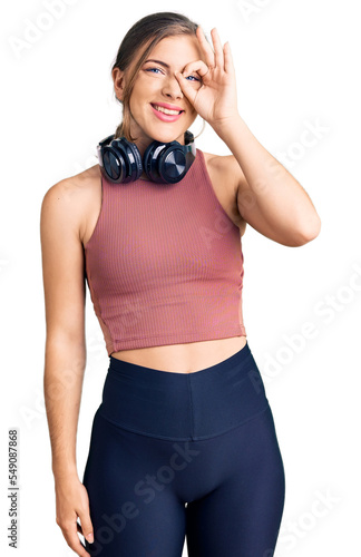 Beautiful caucasian young woman wearing gym clothes and using headphones doing ok gesture with hand smiling, eye looking through fingers with happy face.