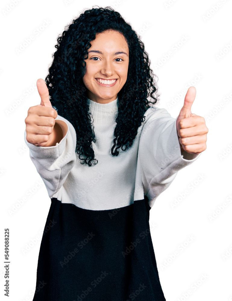 Young hispanic woman with curly hair wearing casual sweater approving doing positive gesture with hand, thumbs up smiling and happy for success. winner gesture. Young hispanic woman with curly hair wearing casual sweater approving doing positive gesture with hand, thumbs up smiling and happy for success. winner gesture.