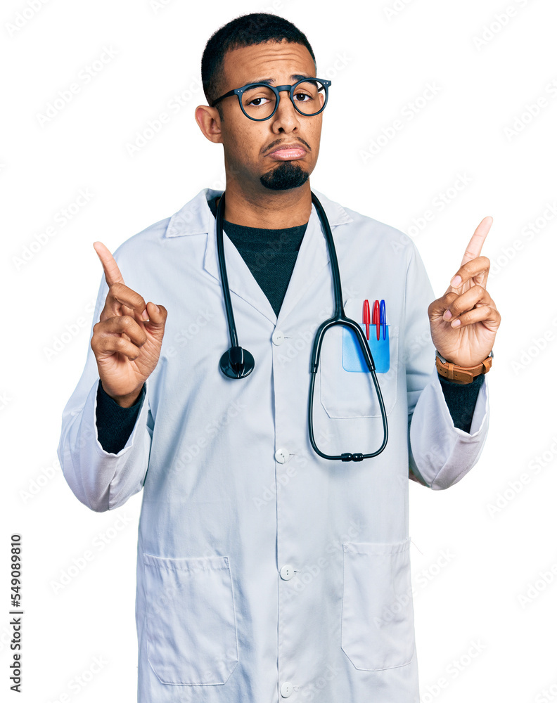 Young african american man wearing doctor uniform and stethoscope pointing up looking sad and upset, indicating direction with fingers, unhappy and depressed. Young african american man wearing doctor uniform and stethoscope pointing up looking sad and upset, indicating direction with fingers, unhappy and depressed.