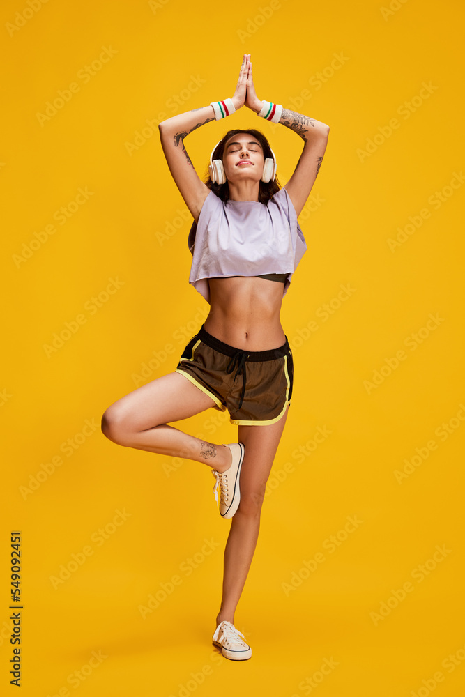 Portrait of young beautiful girl in headphones posing in yoga pose isolated over yellow background. Sportive life - obrazy, fototapety, plakaty 