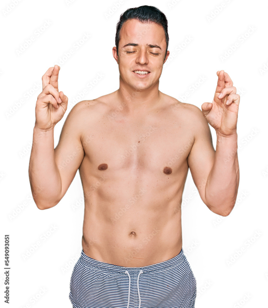 Handsome young man wearing swimwear shirtless gesturing finger crossed smiling with hope and eyes closed. luck and superstitious concept. Handsome young man wearing swimwear shirtless gesturing finger crossed smiling with hope and eyes closed. luck and superstitious concept.