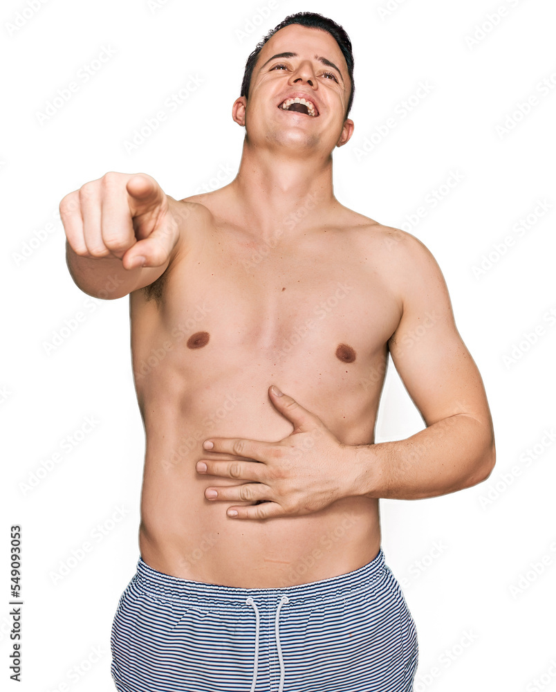 Handsome young man wearing swimwear shirtless laughing at you, pointing finger to the camera with hand over body, shame expression Handsome young man wearing swimwear shirtless laughing at you, pointing finger to the camera with hand over body, shame expression