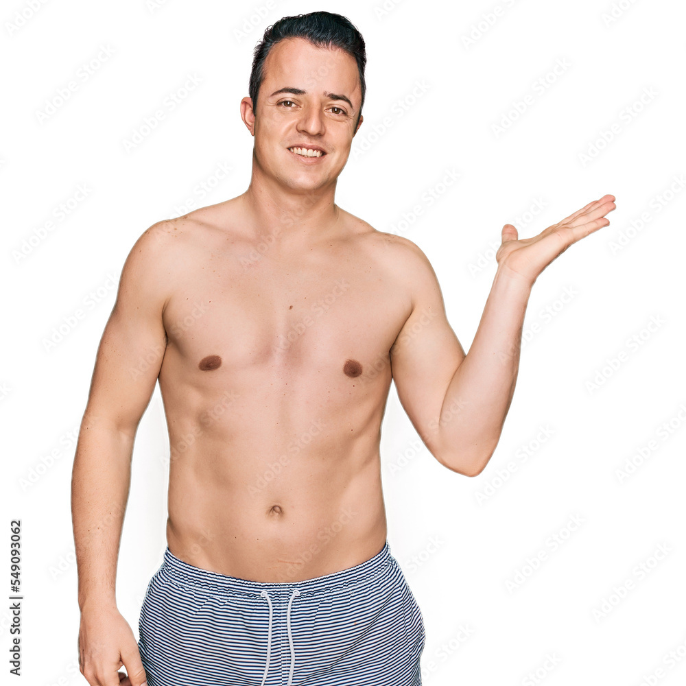 Handsome young man wearing swimwear shirtless smiling cheerful presenting and pointing with palm of hand looking at the camera.