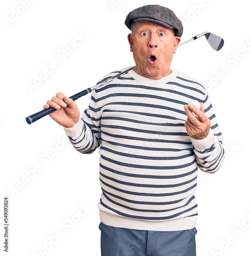 Senior handsome grey-haired man holding golf club and ball scared and amazed with open mouth for surprise, disbelief face