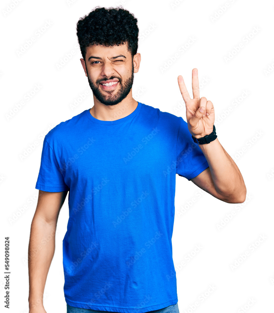 Young arab man with beard wearing casual blue t shirt smiling with happy face winking at the camera doing victory sign with fingers. number two. Young arab man with beard wearing casual blue t shirt smiling with happy face winking at the camera doing victory sign with fingers. number two.