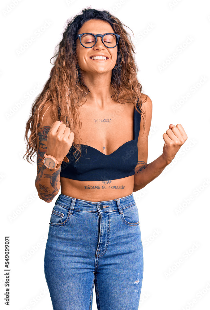 Young hispanic woman with tattoo wearing casual clothes and glasses excited for success with arms raised and eyes closed celebrating victory smiling. winner concept. Young hispanic woman with tattoo wearing casual clothes and glasses excited for success with arms raised and eyes closed celebrating victory smiling. winner concept.