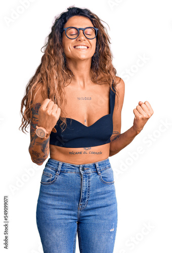 Young hispanic woman with tattoo wearing casual clothes and glasses excited for success with arms raised and eyes closed celebrating victory smiling. winner concept.