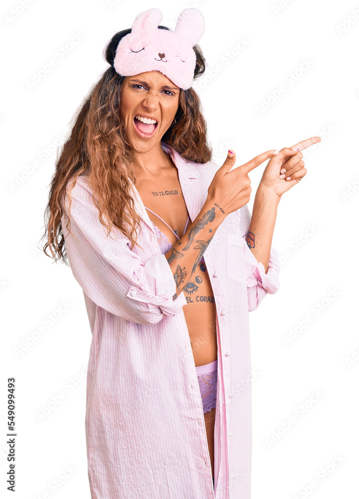Young hispanic woman with tattoo wearing sleep mask and pajama smiling and looking at the camera pointing with two hands and fingers to the side. Young hispanic woman with tattoo wearing sleep mask and pajama smiling and looking at the camera pointing with two hands and fingers to the side.