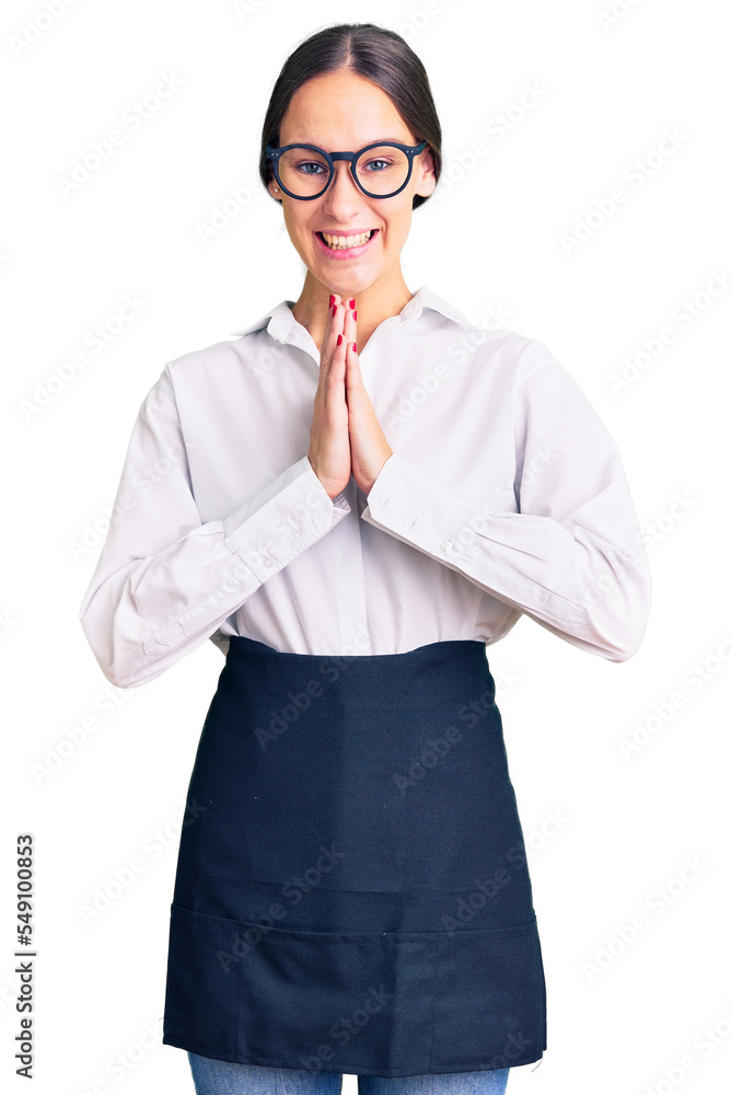 Beautiful brunette young woman wearing professional waitress apron praying with hands together asking for forgiveness smiling confident. Beautiful brunette young woman wearing professional waitress apron praying with hands together asking for forgiveness smiling confident.