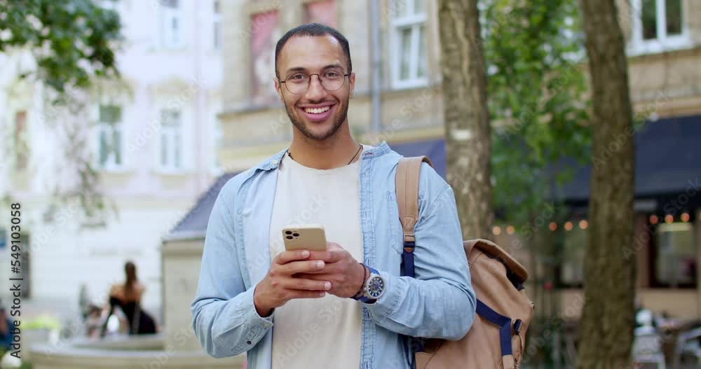 Portrait of cheerful young handsome man in glasses standing at street, texting message on smartphone and smiling to camera. Outdoor. Good-looking male traveler in town chatting and tapping on phone. Portrait of cheerful young handsome man in glasses standing at street, texting message on smartphone and smiling to camera. Outdoor. Good-looking male traveler in town chatting and tapping on phone.