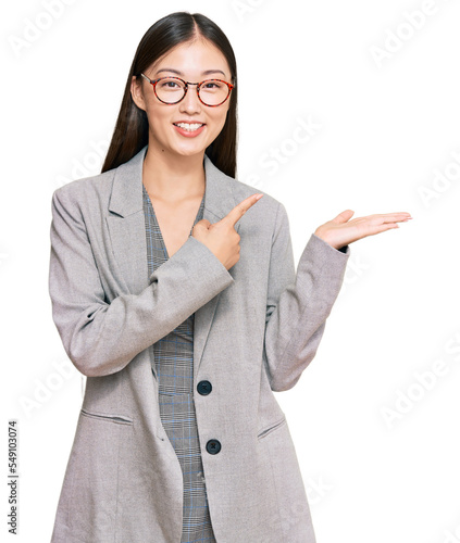 Obraz na plátně Young chinese woman wearing business clothes amazed and smiling to the camera while presenting with hand and pointing with finger