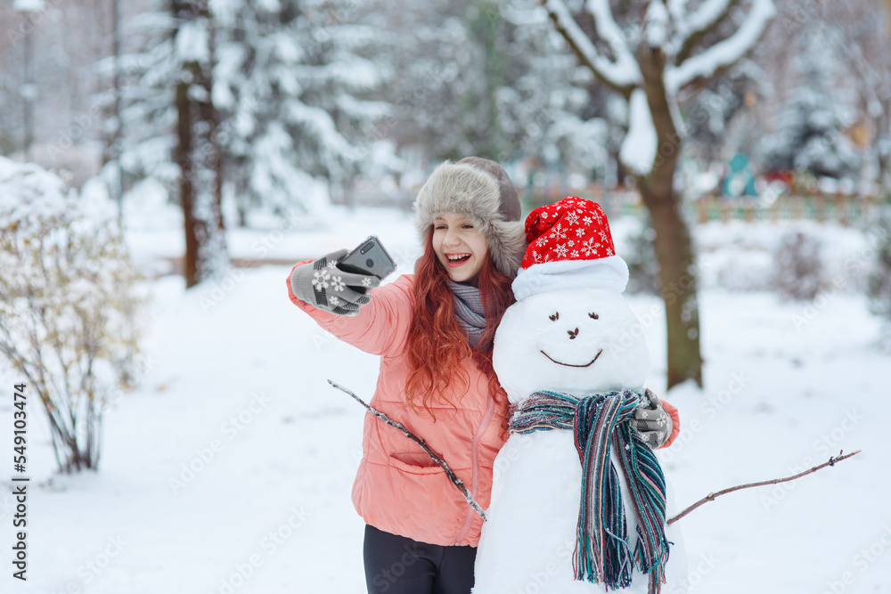 Young happy cheerful laughing girl in fur hat hugging a smiling snowman in christmas hat and scarf, taking selfie with her smartphone in december snow-covered winter park, seasonal  emotional scene
