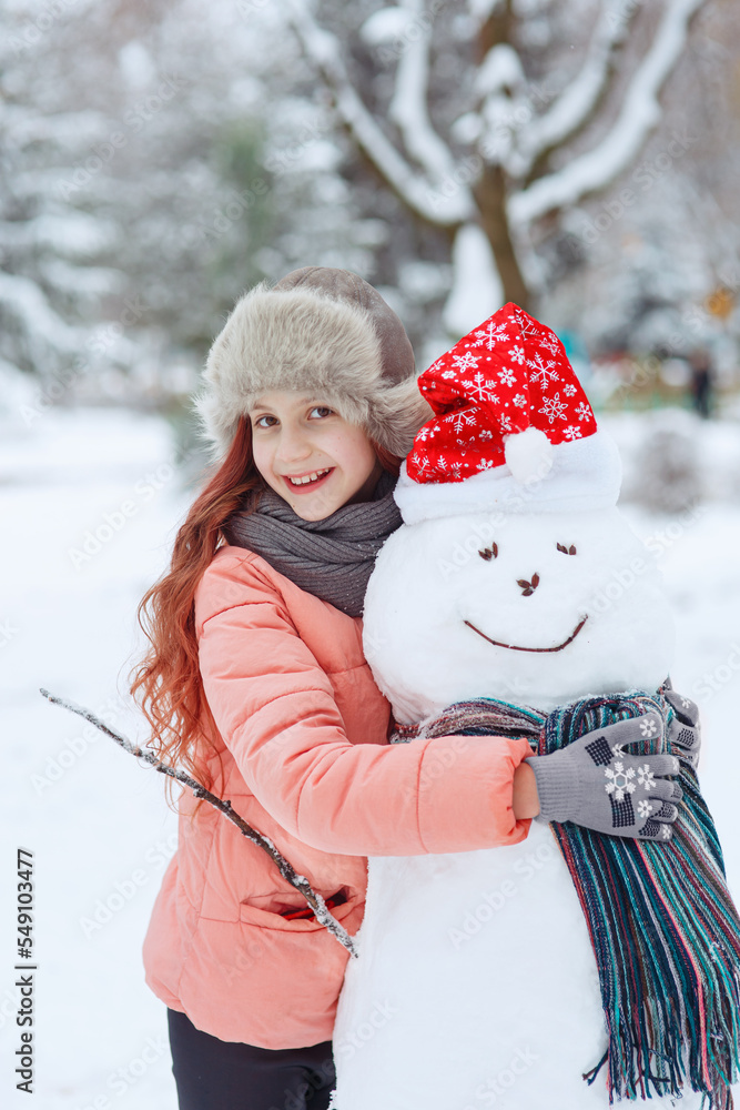 Young cheerful positive girl in fur hat hugging a smiling snowman in christmas hat and scarf and having fun in december snow-covered winter park, seasonal emotional portrait, happiness of childhood Young cheerful positive girl in fur hat hugging a smiling snowman in christmas hat and scarf and having fun in december snow-covered winter park, seasonal emotional portrait, happiness of childhood