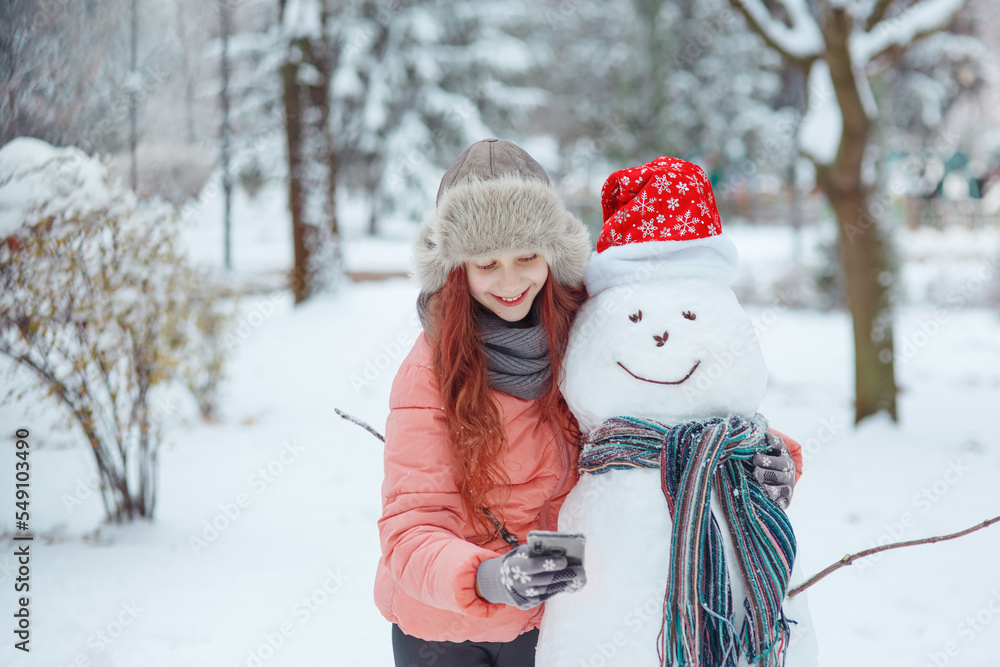 Young happy cheerful laughing girl in fur hat hugging smiling snowman in christmas hat and scarf, showing it her smartphone in december snow-covered winter park, seasonal  emotional outdoor lifestyle