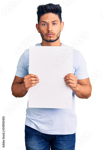 Handsome latin american young man holding cardboard banner with blank space thinking attitude and sober expression looking self confident