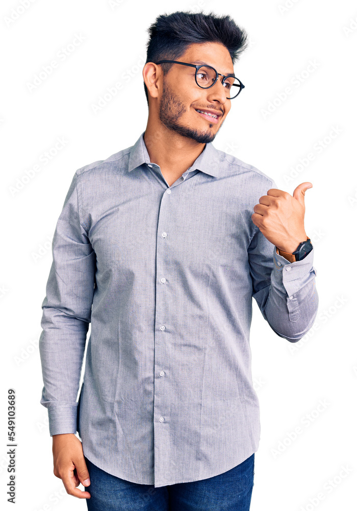 Handsome latin american young man wearing business clothes and glasses smiling with happy face looking and pointing to the side with thumb up. Handsome latin american young man wearing business clothes and glasses smiling with happy face looking and pointing to the side with thumb up.