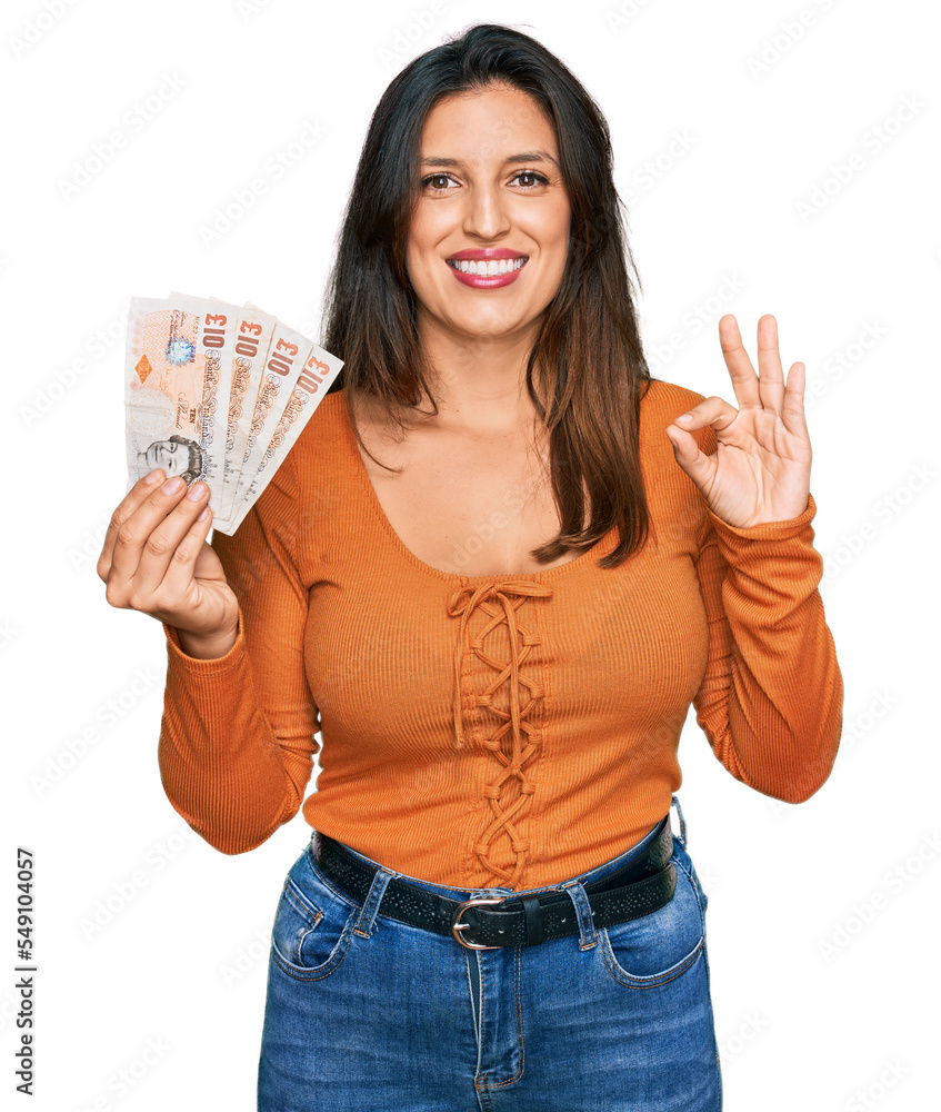 Beautiful hispanic woman holding 10 united kingdom pounds banknotes doing ok sign with fingers, smiling friendly gesturing excellent symbol Beautiful hispanic woman holding 10 united kingdom pounds banknotes doing ok sign with fingers, smiling friendly gesturing excellent symbol