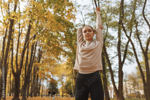Low angle view of sporty woman stretching raised arms and exercising during fitness training in autumn park on sunny day 