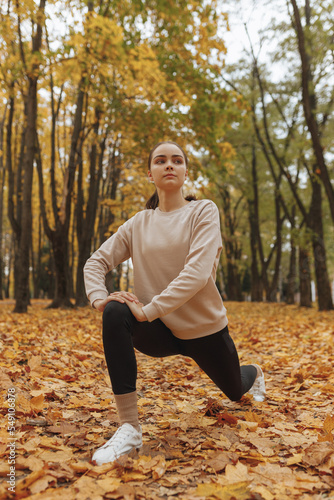 Full length of flexible female runner stretching legs and doing lunge exercise while training in autumn park and looking away 