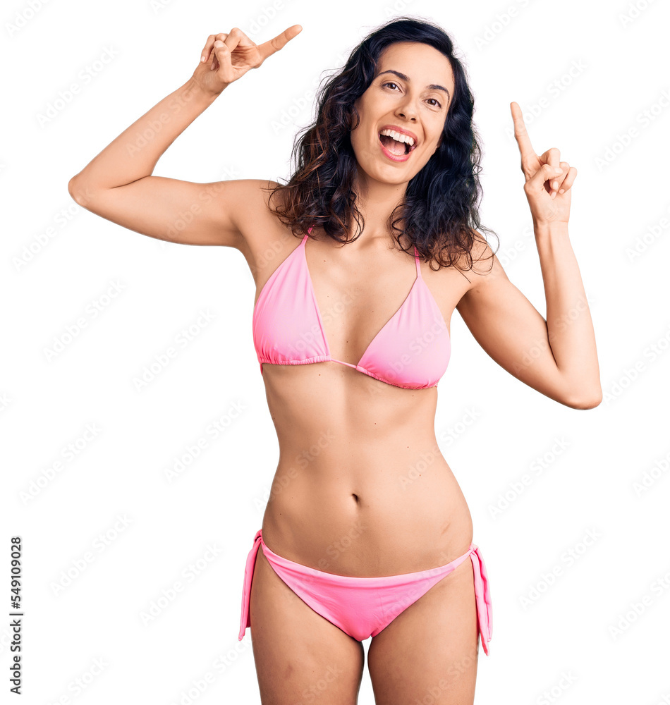 Young beautiful hispanic woman wearing bikini smiling amazed and surprised and pointing up with fingers and raised arms. Young beautiful hispanic woman wearing bikini smiling amazed and surprised and pointing up with fingers and raised arms.