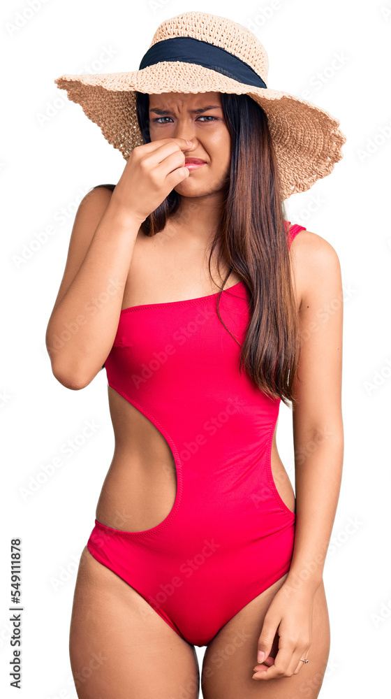 Young beautiful latin girl wearing swimwear and summer hat smelling something stinky and disgusting, intolerable smell, holding breath with fingers on nose. bad smell Young beautiful latin girl wearing swimwear and summer hat smelling something stinky and disgusting, intolerable smell, holding breath with fingers on nose. bad smell