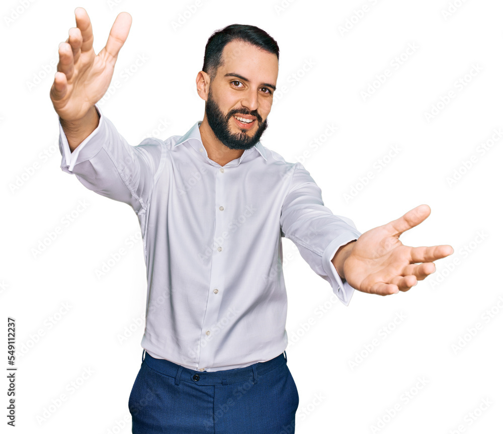 Naklejka premium Young man with beard wearing business shirt looking at the camera smiling with open arms for hug. cheerful expression embracing happiness.