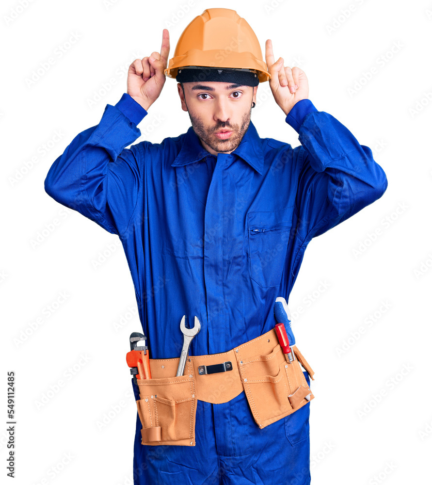 Young hispanic man wearing worker uniform doing funny gesture with finger over head as bull horns Young hispanic man wearing worker uniform doing funny gesture with finger over head as bull horns