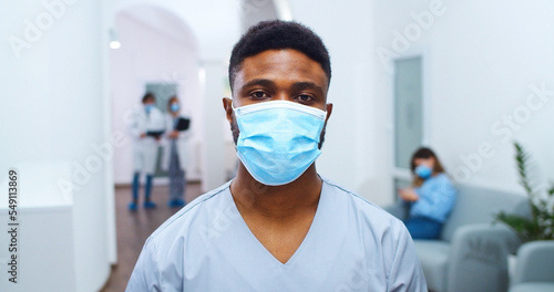 Close up portrait of African American young male doctor in medical mask standing in hospital hall and looking at camera. Mixed-races healthcare professionals on background Coronavirus epidemic concept