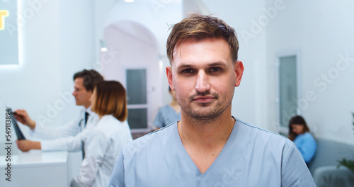 Close up portrait of handsome Caucasian young happy male doctor surgeon standing in hospital hall and looking at camera in good mood. Multi-ethnic healthcare professionals on background