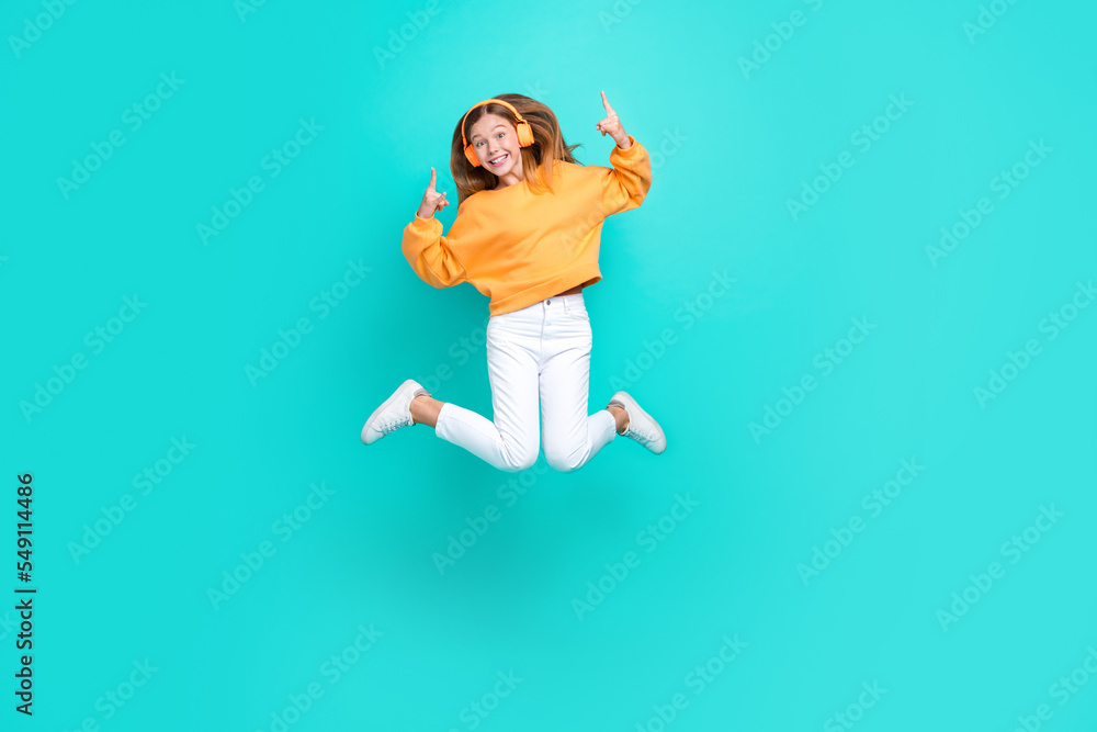 Full length photo of cool funky school girl dressed orange hoodie jumping high showing hard rock listening songs isolated teal color background Full length photo of cool funky school girl dressed orange hoodie jumping high showing hard rock listening songs isolated teal color background