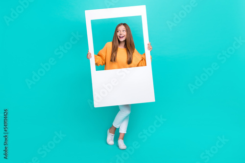 Full length photo of adorable dreamy school girl dressed orange hoodie tacking photo looking empty space isolated teal color background