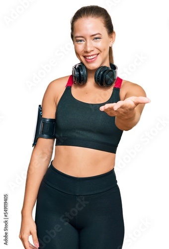 Beautiful blonde woman wearing gym clothes and using headphones smiling friendly offering handshake as greeting and welcoming. successful business.
