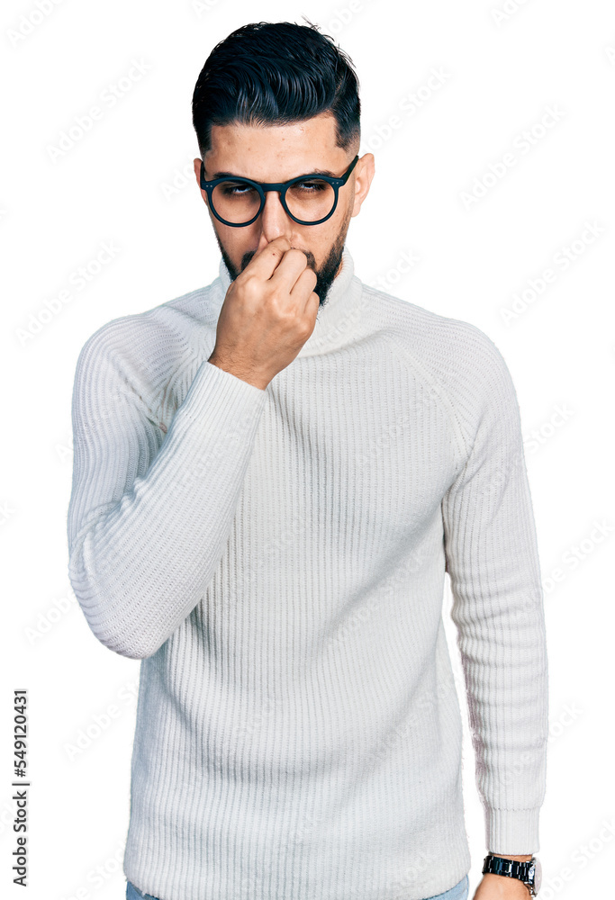 Young arab man with beard wearing elegant turtleneck sweater and glasses smelling something stinky and disgusting, intolerable smell, holding breath with fingers on nose. bad smell Young arab man with beard wearing elegant turtleneck sweater and glasses smelling something stinky and disgusting, intolerable smell, holding breath with fingers on nose. bad smell