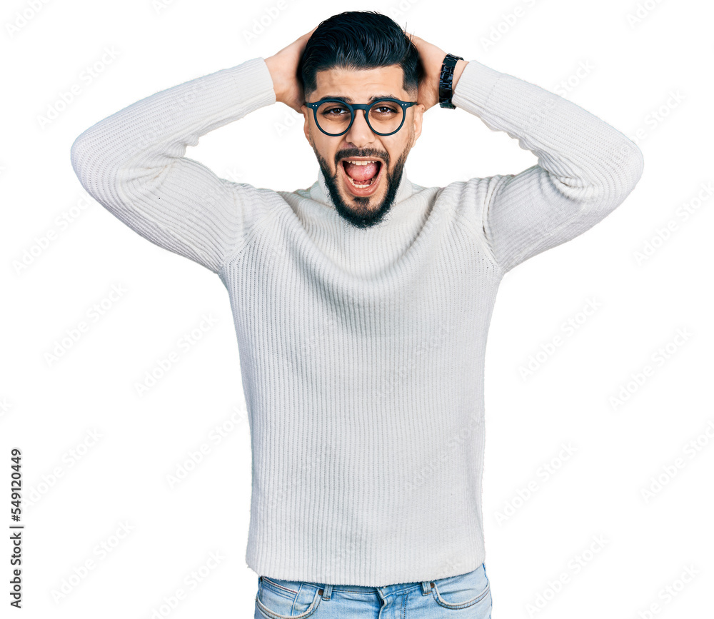 Young arab man with beard wearing elegant turtleneck sweater and glasses crazy and scared with hands on head, afraid and surprised of shock with open mouth Young arab man with beard wearing elegant turtleneck sweater and glasses crazy and scared with hands on head, afraid and surprised of shock with open mouth