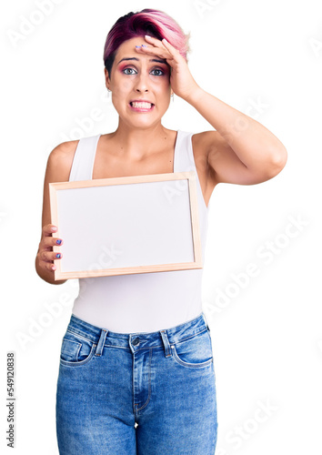 Young beautiful woman with pink hair holding empty white chalkboard stressed and frustrated with hand on head, surprised and angry face