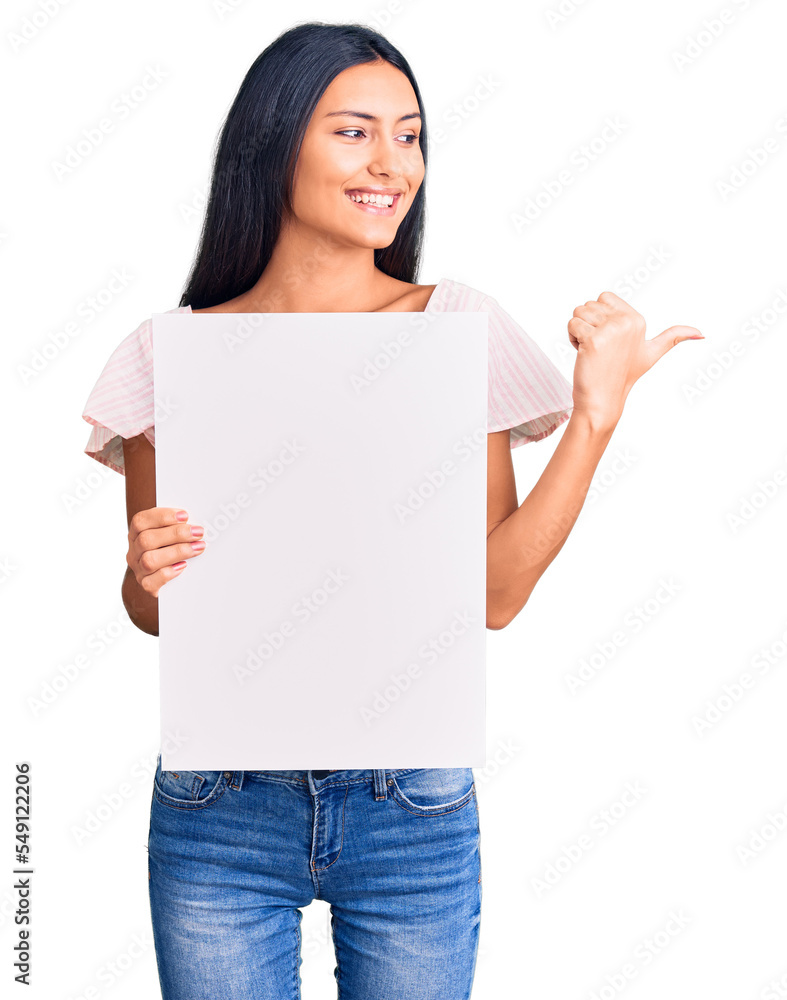 Young beautiful latin girl holding blank empty banner pointing thumb up to the side smiling happy with open mouth Young beautiful latin girl holding blank empty banner pointing thumb up to the side smiling happy with open mouth