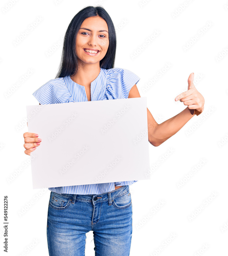 Young beautiful latin girl holding blank empty banner smiling happy pointing with hand and finger Young beautiful latin girl holding blank empty banner smiling happy pointing with hand and finger