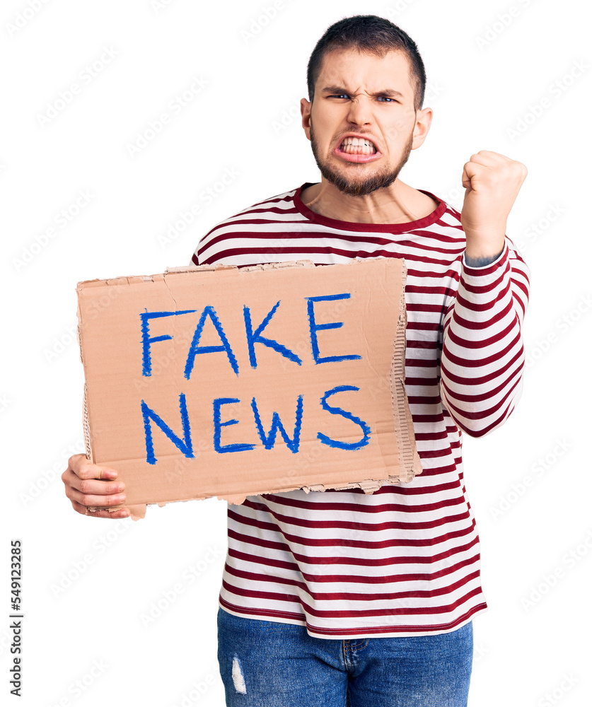 Young handsome man holding fake news message banner annoyed and frustrated shouting with anger, yelling crazy with anger and hand raised Young handsome man holding fake news message banner annoyed and frustrated shouting with anger, yelling crazy with anger and hand raised