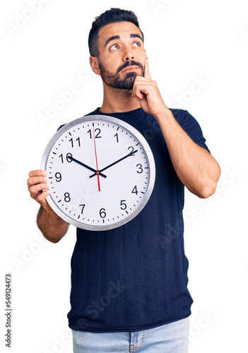 Young hispanic man holding big clock serious face thinking about question with hand on chin, thoughtful about confusing idea
