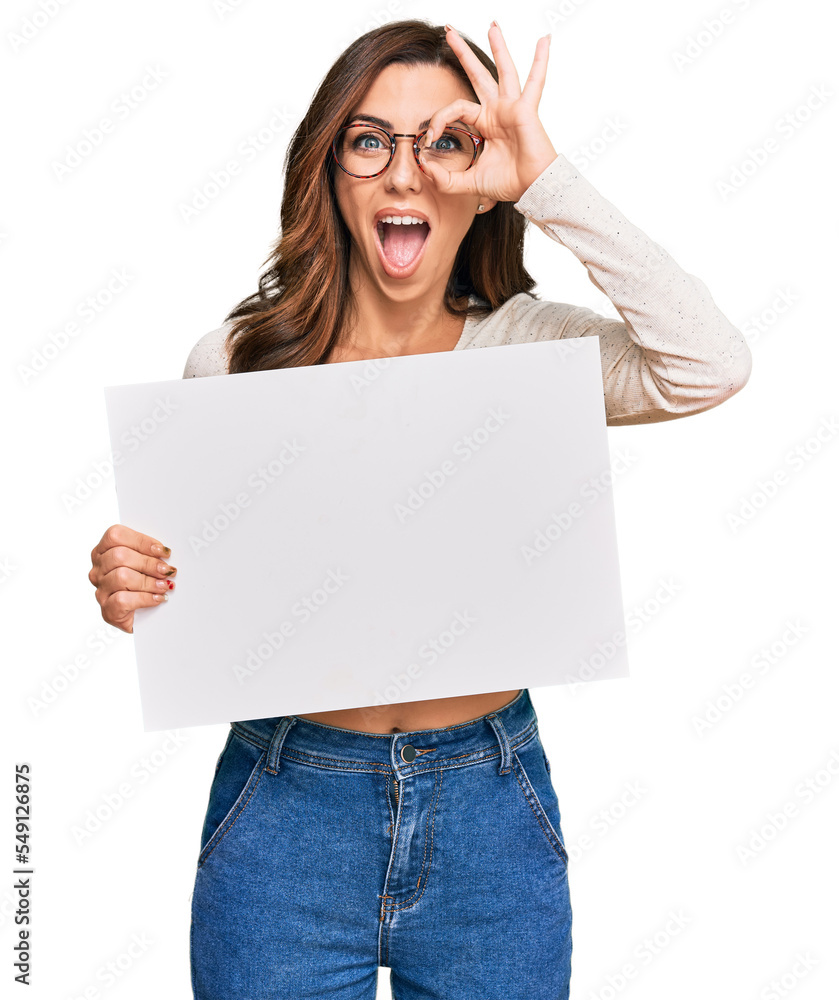 Young brunette woman holding blank empty banner smiling happy doing ok sign with hand on eye looking through fingers Young brunette woman holding blank empty banner smiling happy doing ok sign with hand on eye looking through fingers