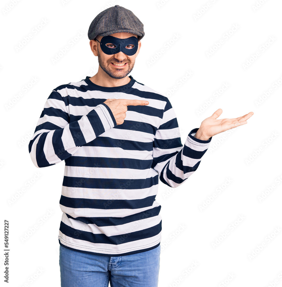 Young handsome man wearing burglar mask amazed and smiling to the camera while presenting with hand and pointing with finger. Young handsome man wearing burglar mask amazed and smiling to the camera while presenting with hand and pointing with finger.