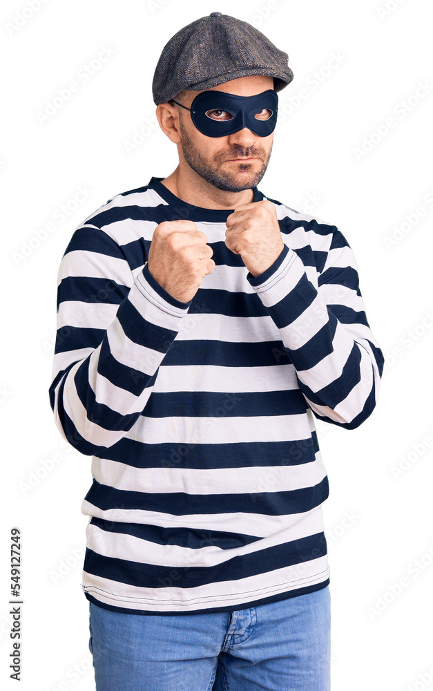 Young handsome man wearing burglar mask ready to fight with fist defense gesture, angry and upset face, afraid of problem Young handsome man wearing burglar mask ready to fight with fist defense gesture, angry and upset face, afraid of problem
