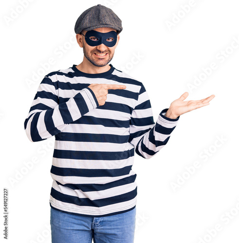 Young handsome man wearing burglar mask amazed and smiling to the camera while presenting with hand and pointing with finger.