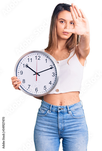 Young beautiful girl holding big clock with open hand doing stop sign with serious and confident expression, defense gesture